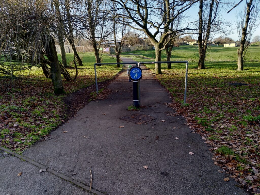 An entrance to Landseer Park, Ipswich which has a bollard with blue cycle/pedestrians shared use sign. Just beyond the bollard are 2 barriers which are difficult to get a bike past. To the left of the barriers is a desire line to avoid the difficult to navigate barriers.