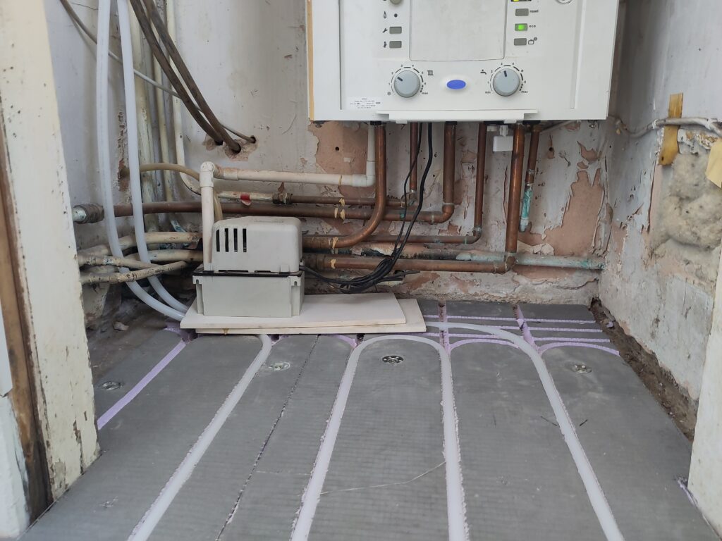 Insulated backer board on the floor with pipes in the grooves, and gas boiler in the background on the wall.