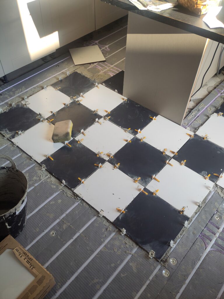 16 black and white tiles on the floor above the insulated backer board. Pack of white tiles and pail of adhesive just in view.