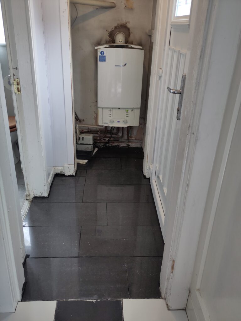 View of small room by the back door with the dark drey granite floor tiles. Gas boiler on back wall, door to outside on right, utility room to the left. Kitchen and cupboard doors removed.