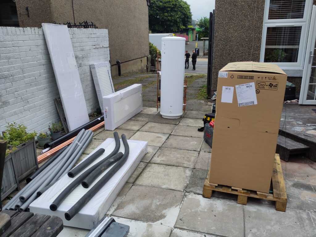Back garden with the new equipment. Heat pump in cardboard package. Now hot water tank, radiators, and other pipework.