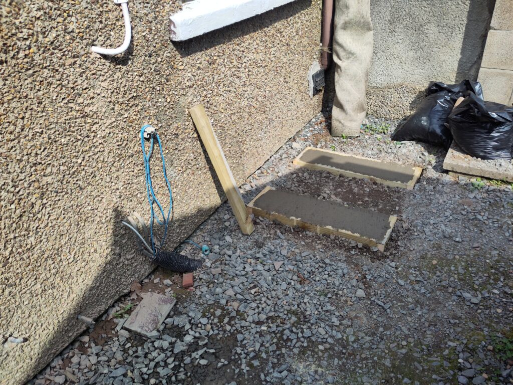 Concrete slabs where the heat pump will sit. Concrete curing. By the back wall of the house. Can see a window sill just in shot at the top of the picture.