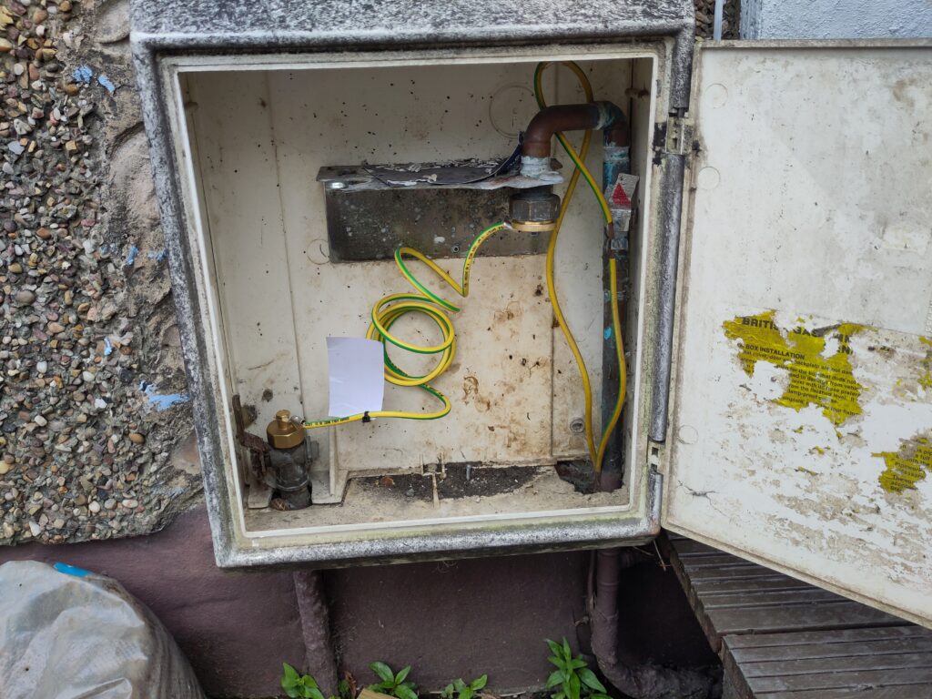 Gas meter box with the meter removed. There is a yellow and green earthing cable linking the two pipes.